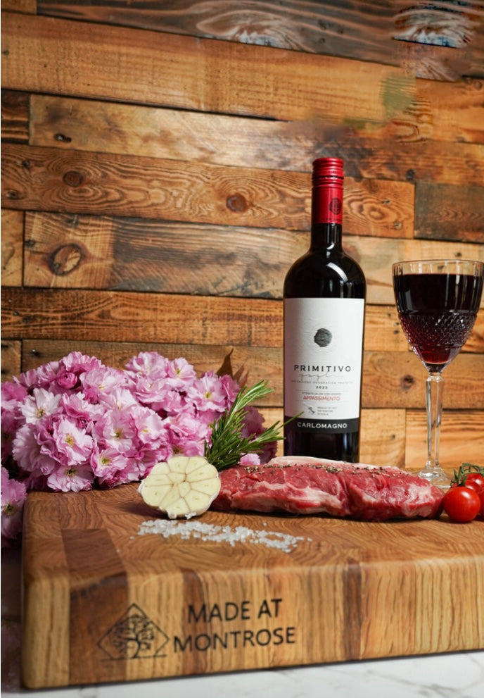 Portrait view of the Montrose Edition solid oak end grain chopping board with engraved Made at Montrose logo, steak, garlic and red wine.