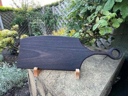 Angled view of a handmade dark oak charcuterie board showing its circular handle, natural wood grain and hand-torched finish 