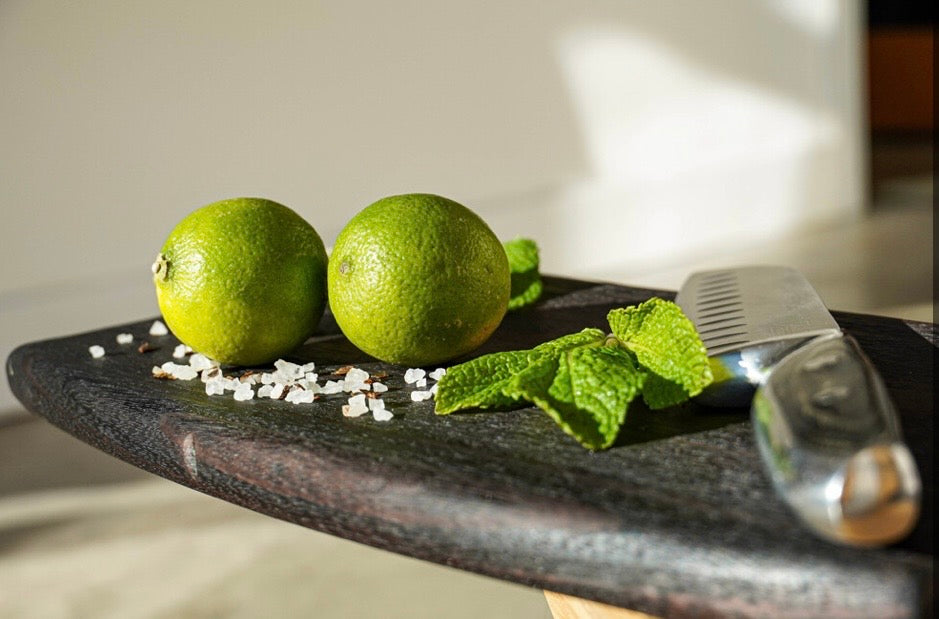 Handmade dark oak charcuterie board laid flat with fresh limes, mint leaves, sea salt and a kitchen knife.