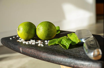 Handmade dark oak charcuterie board laid flat with fresh limes, mint leaves, sea salt and a kitchen knife.
