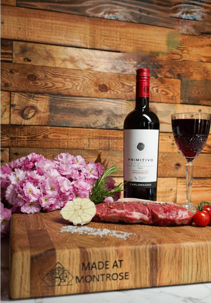Portrait view of the Montrose Edition solid oak end grain chopping board with engraved Made at Montrose logo, steak, garlic and red wine.